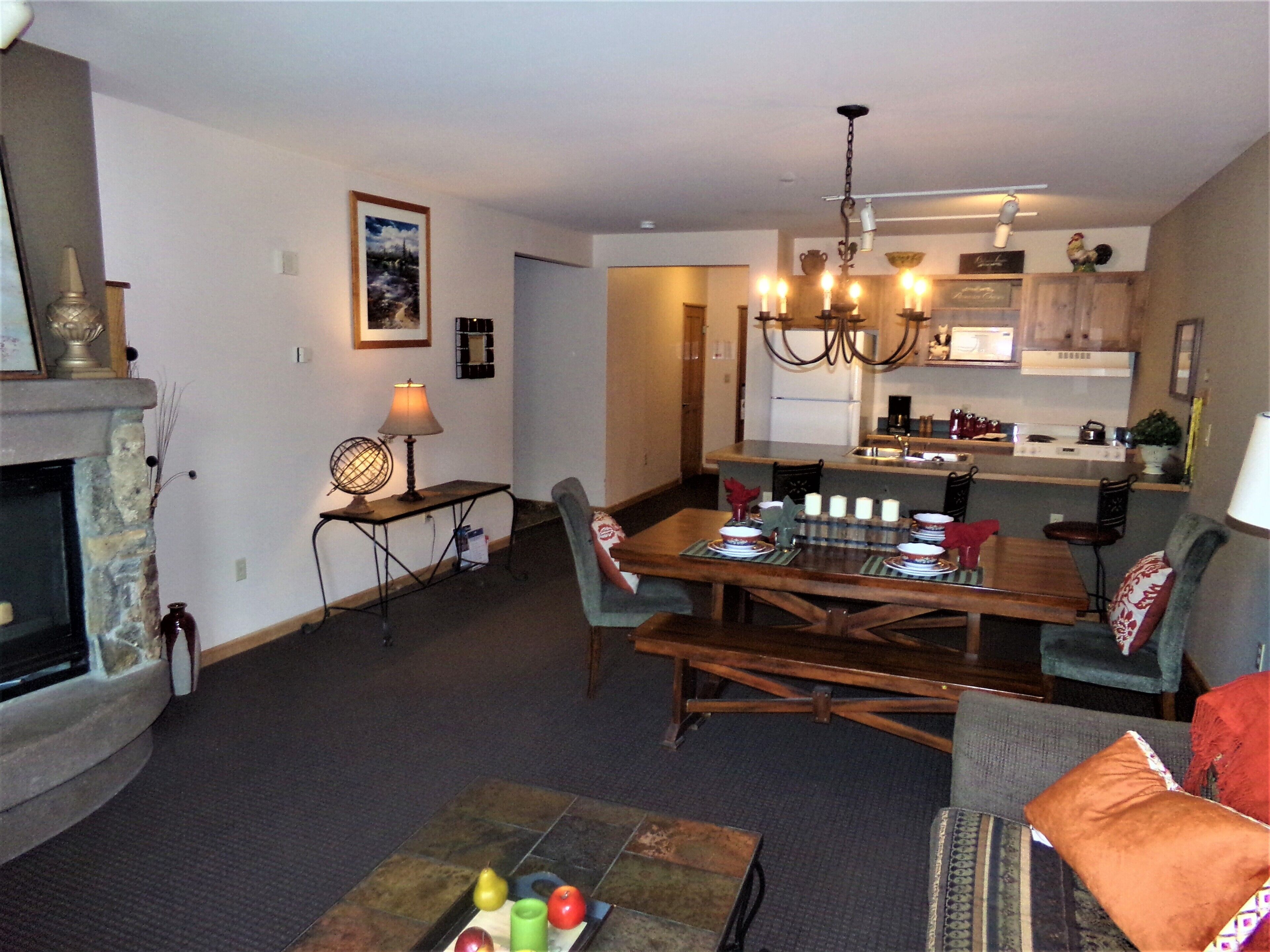 A dining area with a wooden table set for four, a fireplace, and a kitchen in the background.