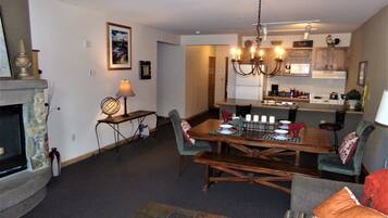 A dining area with a wooden table set for four, a fireplace, and a kitchen in the background.