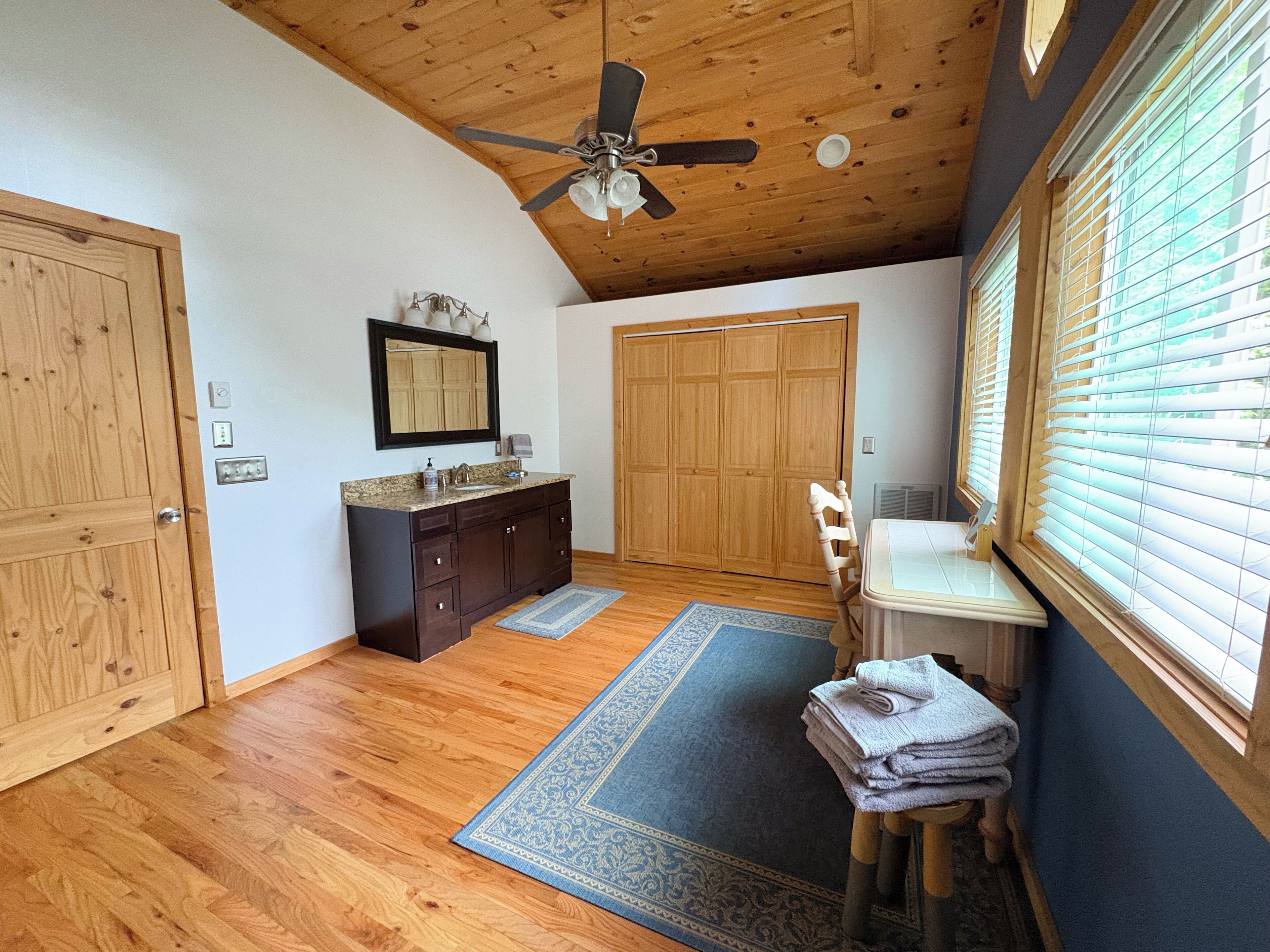A room with wooden floors, a ceiling fan, a vanity with a mirror, a wooden door, and a window with blinds.