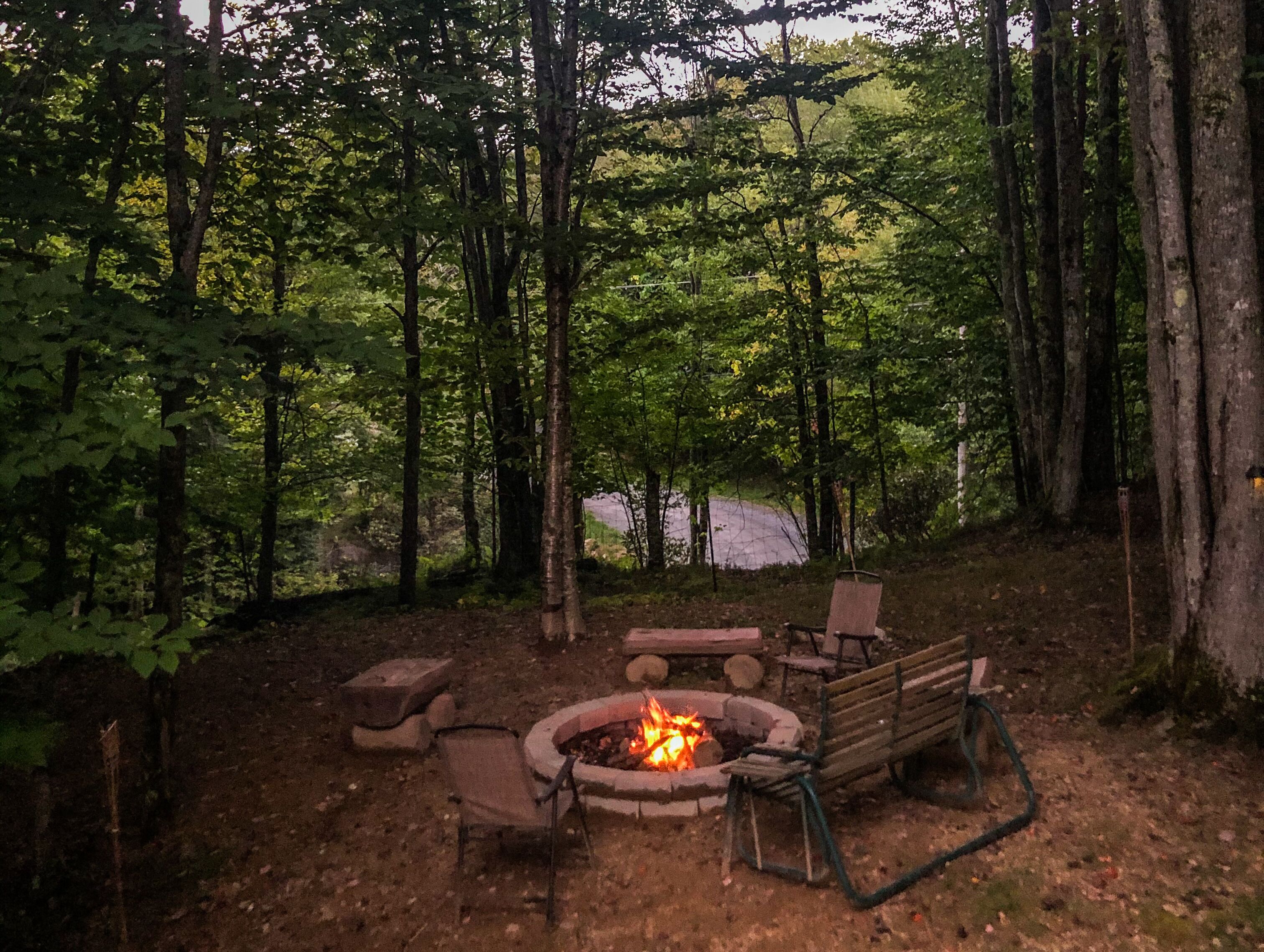Un terrain de camping situé dans une forêt, équipé d’un foyer, de bancs et d’une chaise.