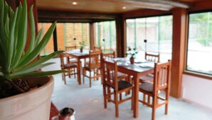 A restaurant with wooden tables and chairs, large windows, and a potted plant in the foreground. - Morada Fenix Hospedagem (Palhoça)