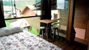 A bedroom with a bed, a wooden desk, two chairs, and a large window overlooking a village. - Morada Fenix Hospedagem (Palhoça)