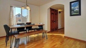 Un comedor con una mesa de madera, sillas negras y un banco. Se ve una ventana con cortinas, una puerta de madera y un cuadro en la pared.