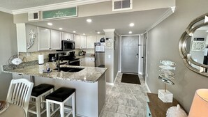 A modern kitchen with white cabinets, a granite countertop, and stainless steel appliances. There are two bar stools and a decorative fish wall art piece. - 507 "A" Relaxing Bliss! Sept 13-20 Open! Book Soon! (Gulf Shroes)