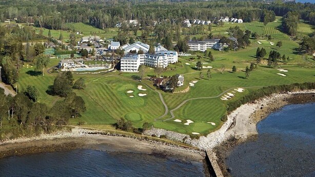 Un parcours de golf doté de plusieurs bâtiments, dont un grand hôtel et plusieurs structures plus petites, le tout entouré d’une végétation luxuriante et bordé d’un plan d’eau.