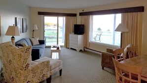 Une chambre d’hôtel équipée d’un canapé, d’un fauteuil, d’une table à manger et de chaises. On y trouve une télévision posée sur une commode, ainsi qu’une fenêtre avec des rideaux.