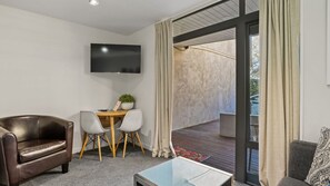 A modern living room with a leather sofa, a wooden dining table with two chairs, a flat-screen TV mounted on the wall, and a glass coffee table. - The Alps Studio - Cosy one bed right in the centre of Queenstown (Queenstown)
