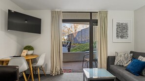 A modern living room with a flat-screen TV mounted on the wall, a sofa with patterned cushions, a coffee table, and a dining area with a table and chairs. - The Alps Studio - Cosy one bed right in the centre of Queenstown (Queenstown)