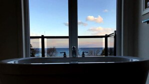 Een vrijstaand bad met uitzicht op zee, zichtbaar door het balkonhek.