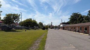 A motel with a parking lot, a gas station in the background, and a grassy area with trees. - Studio 1 Motel (Cobourg)