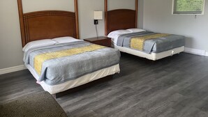 Hotelkamer met twee bedden, houten hoofdborden, een nachtkastje, een lamp en een raam met jaloezieën.
