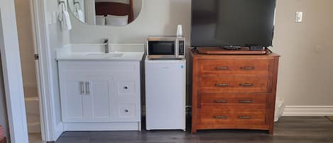 Une pièce avec une coiffeuse blanche, un four à micro-ondes, un téléviseur posé sur une commode en bois et un miroir au mur.