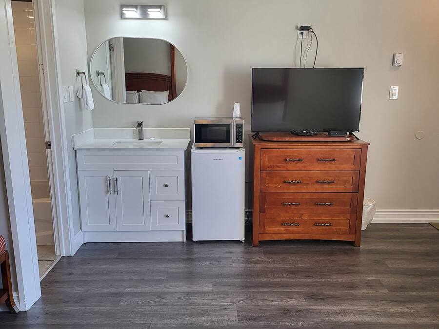 A room with a white vanity, a microwave, a television on a wooden dresser, and a mirror on the wall.