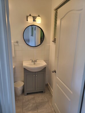 Une salle de bain avec un lavabo blanc, un miroir rond et une porte avec une fenêtre.