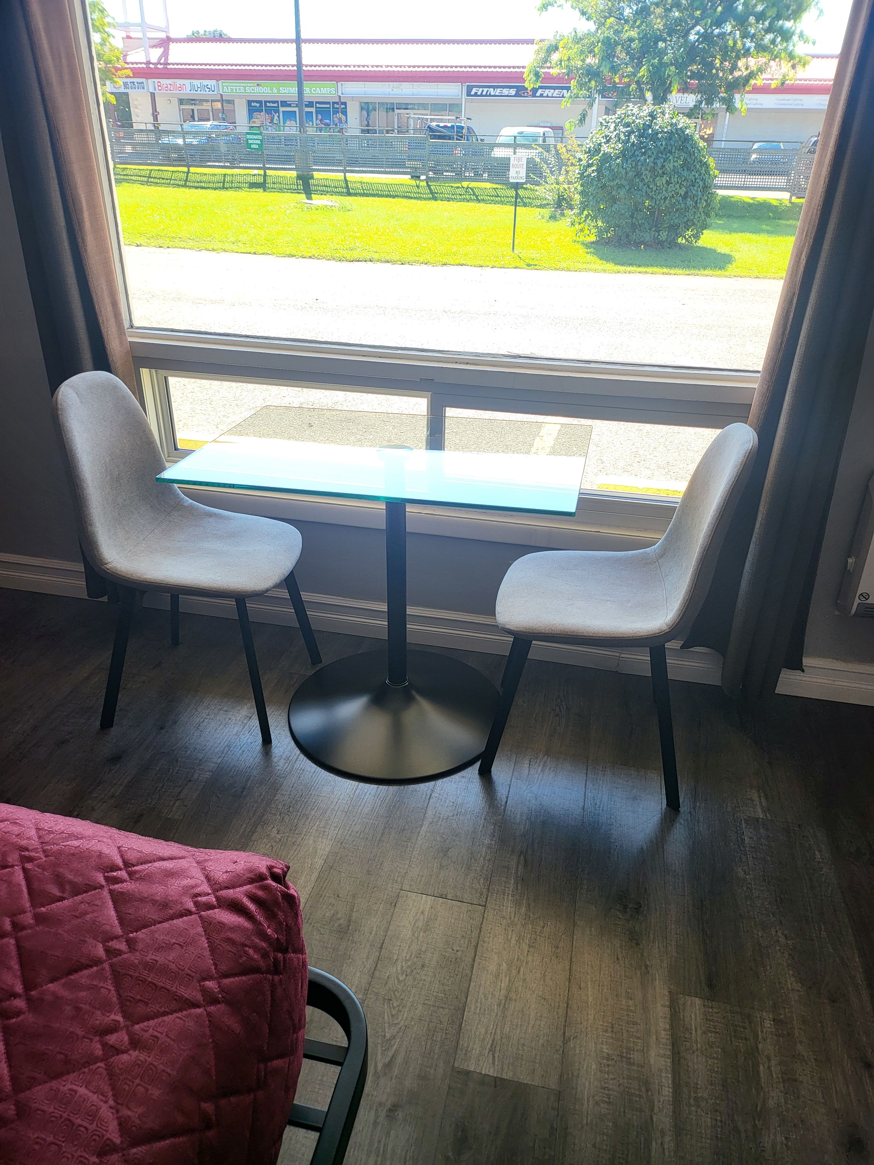Een kamer met uitzicht op een parkeerplaats en een gebouw met borden. Er staat een tafel met twee stoelen en een rode deken op de vloer.