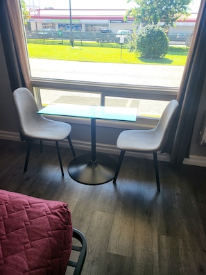 A room with a window view of a parking lot and a building with signage. There is a table with two chairs and a red blanket on the floor. - Studio 1 Motel (Cobourg)