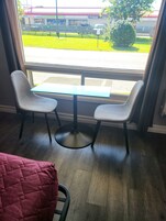 Una habitación con ventana que ofrece vista a un estacionamiento y un edificio con señalización. Hay una mesa con dos sillas y una manta roja en el suelo.