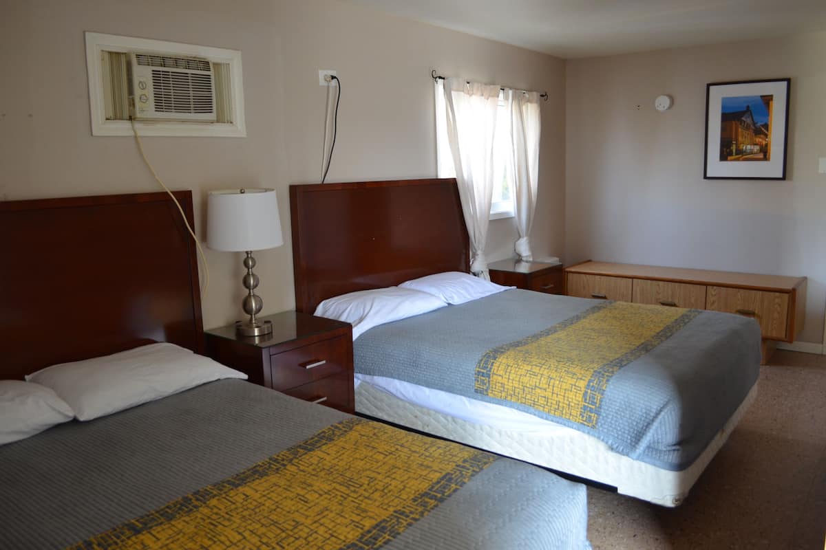 Habitación de hotel con dos camas, una mesita de noche con lámpara, una ventana con cortinas, aire acondicionado instalado en la pared y un cuadro enmarcado.