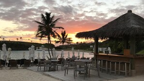 A beachside restaurant with outdoor seating, a thatched-roof structure, and a view of the sunset over the water. - PalmSprings Accommodations - 2 Bedroom Apt., Near Horseshoe Bay (Southampton Parish)
