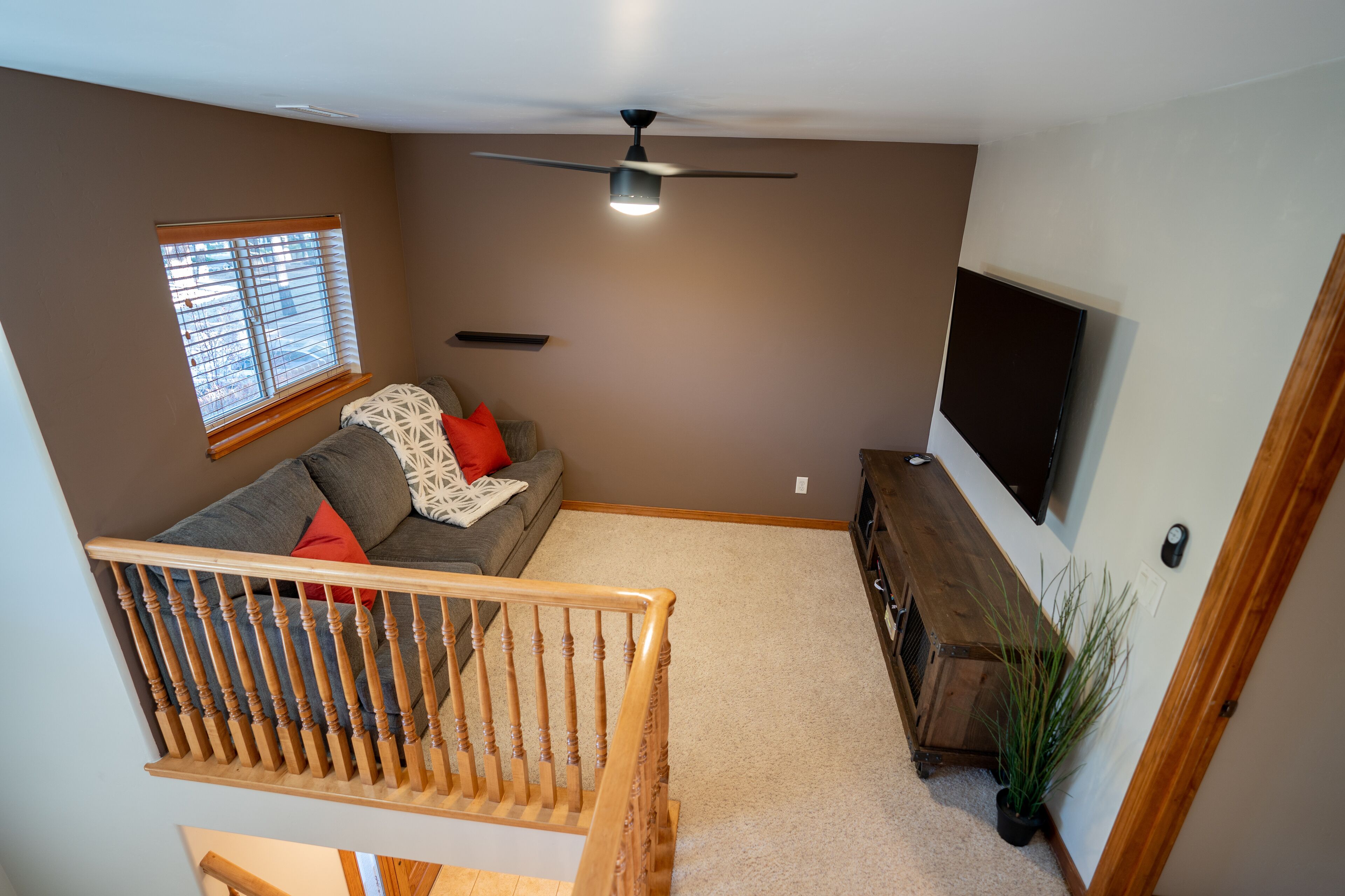 Un salon avec un escalier en bois, un canapé gris, un ventilateur de plafond et un téléviseur fixé au mur.