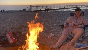 A beach scene with a campfire, a person sitting on a beach chair, and a pier in the background.