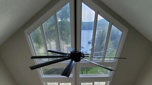 A spacious room with a high ceiling, a ceiling fan, a large window with a view of a lake, a wooden dining table, and a sofa. - Lakefront Home in Private Cove of Deerfield Resort with Boat Slip. Pet-Friendly! (LaFollette)