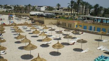 A beach with numerous thatched umbrellas and lounge chairs, a clear sky, and buildings in the background.