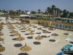 A beach with numerous thatched umbrellas and lounge chairs, a clear sky, and buildings in the background.