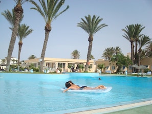 A swimming pool with palm trees, lounge chairs, and a building in the background. - Les Palmiers (Monastir)