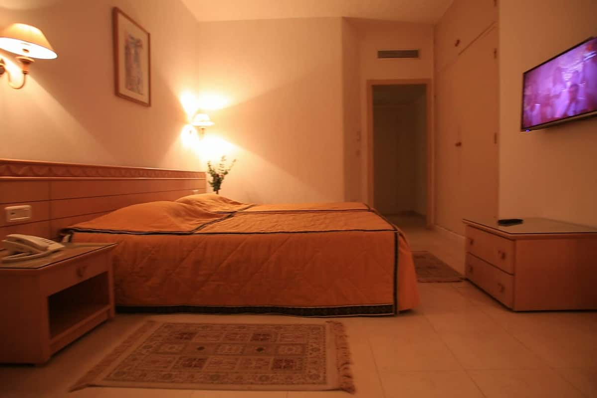 A hotel room with a bed, bedside table, lamp, TV, and a small cabinet.