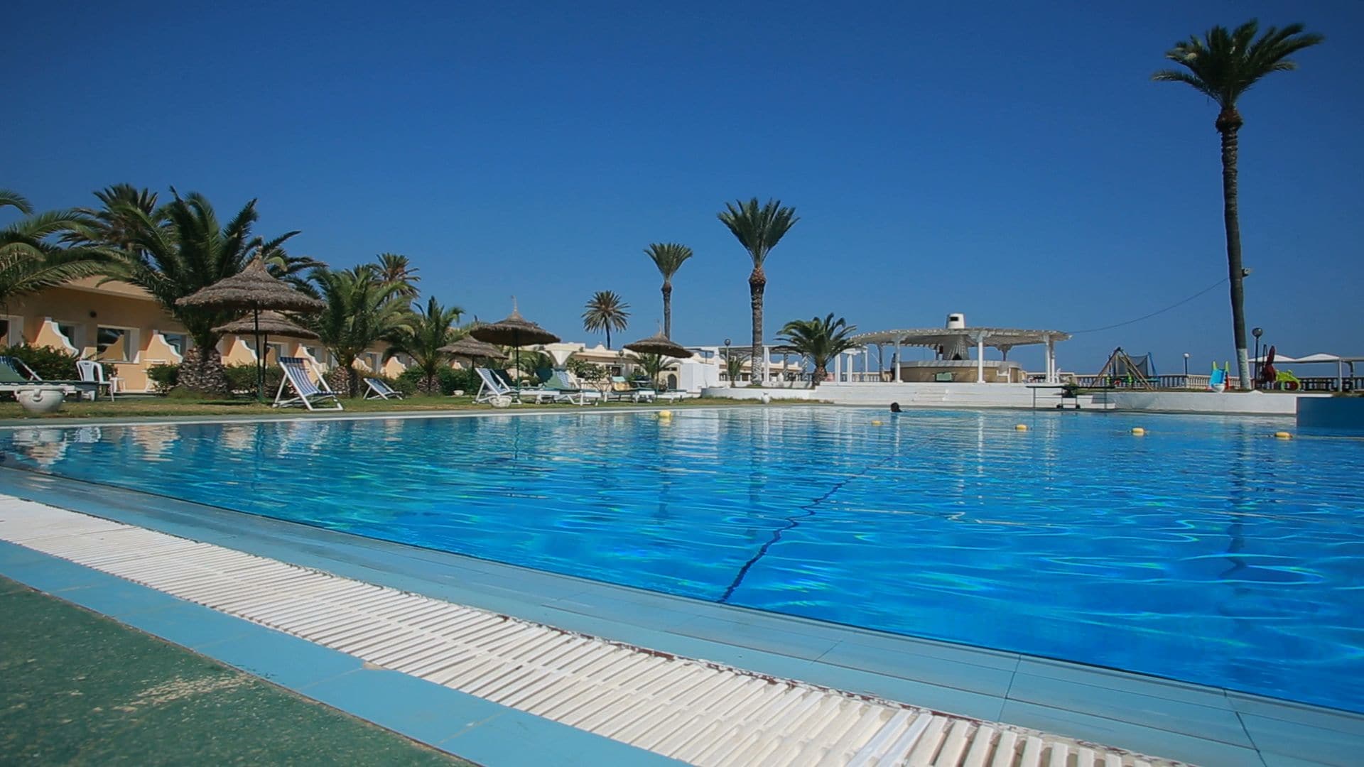 Piscina con sillas de descanso y palmeras, rodeada de edificios con techos de paja.