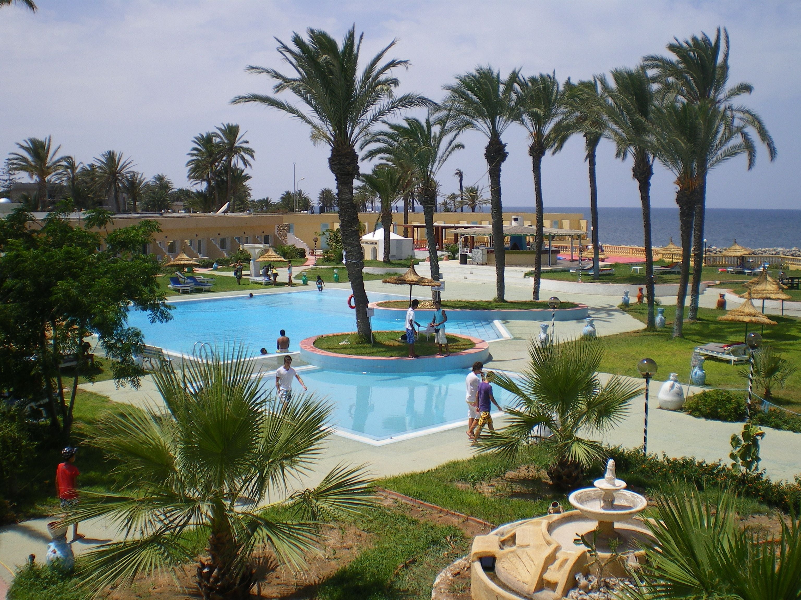Een resort met een centraal gelegen zwembad, palmbomen en uitzicht op zee.