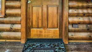 Entrada a una cabaña de madera con una puerta, una corona decorativa, dos faroles y una placa de metal que dice "WHISPERSING PINES".