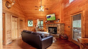 Interior de una cabaña de troncos con chimenea, un sofá de cuero, un televisor y una cabeza de ciervo montada en la pared.
