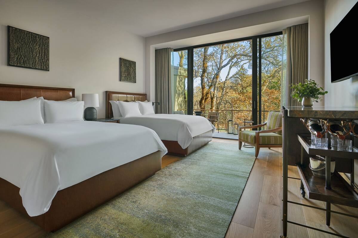 A hotel room with two beds, a balcony with a view of trees, and a dining area with a table and chairs.