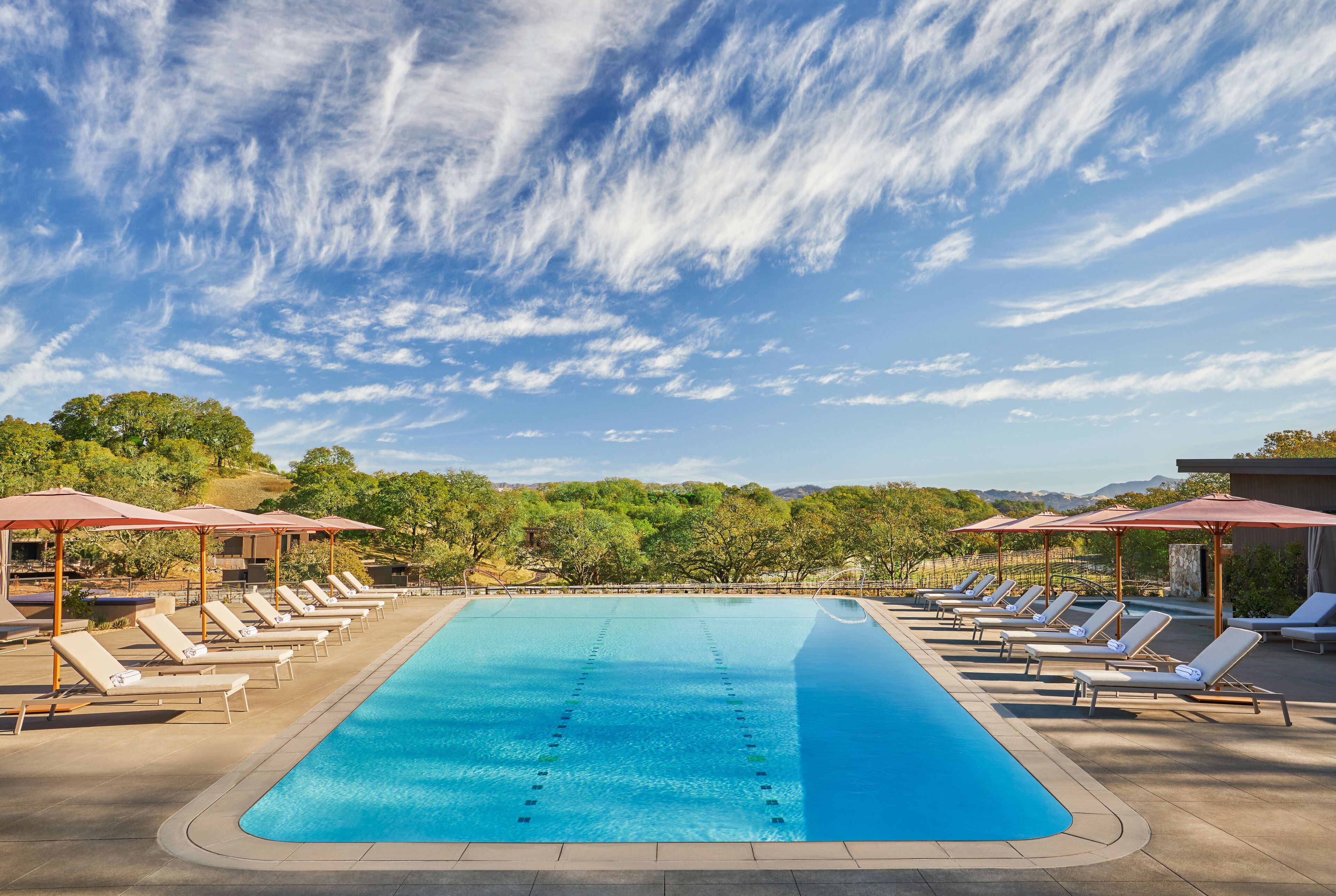 Una piscina con sillas de descanso y sombrillas, rodeada de árboles y un cielo despejado.
