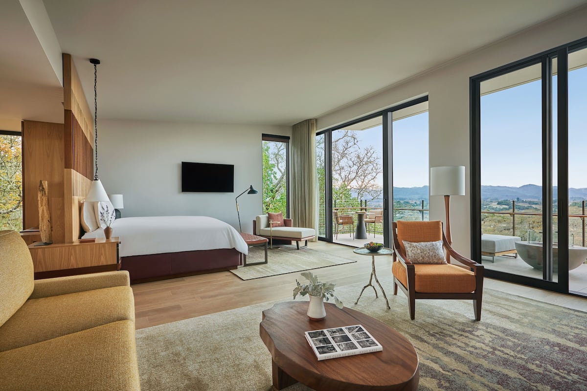 A modern hotel room with a large bed, a sofa, a coffee table, and a balcony with a view of the countryside.