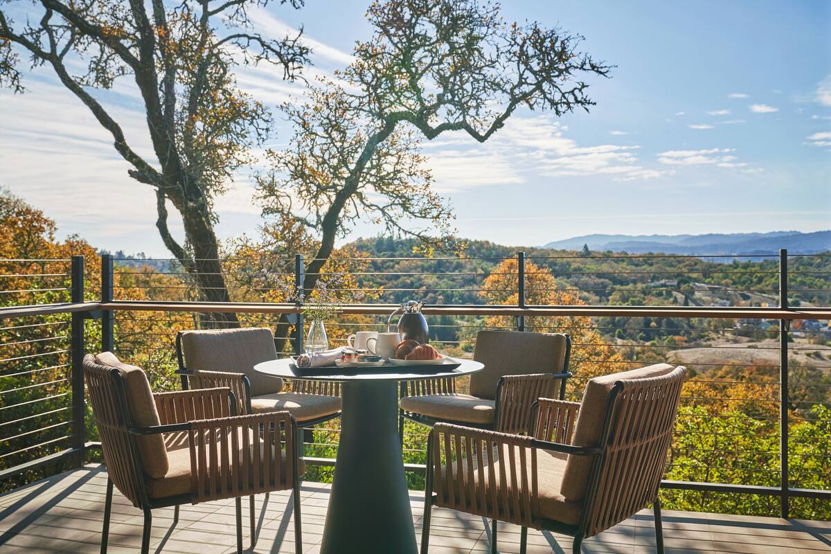A terrace with wicker chairs and a table, overlooking a scenic landscape with trees and distant hills.