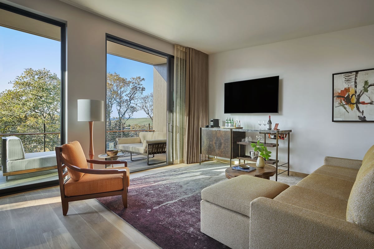 A modern living room with a sofa, armchair, and a coffee table. There is a TV mounted on the wall and a window with a view of trees.