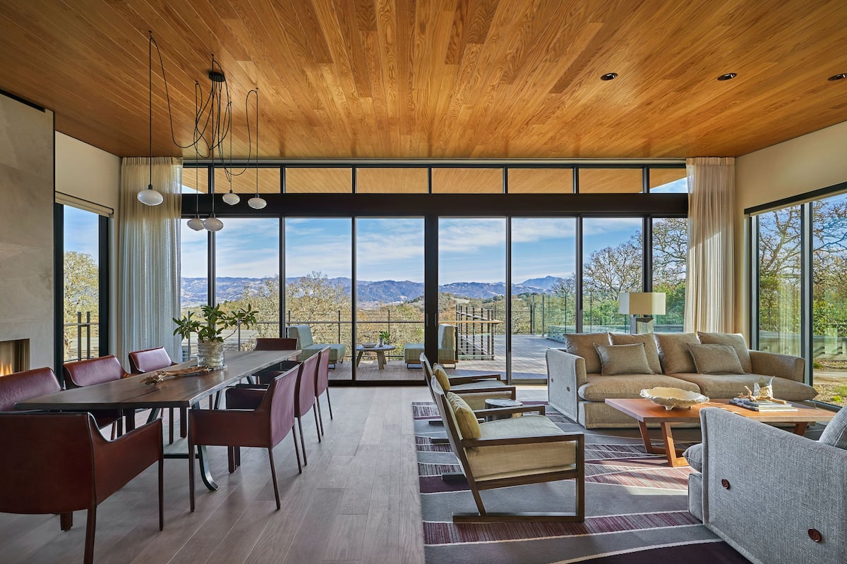 A spacious living area with a wooden ceiling, large windows offering a view of the mountains, a dining area with a long table and chairs, and a seating area with sofas and a coffee table.