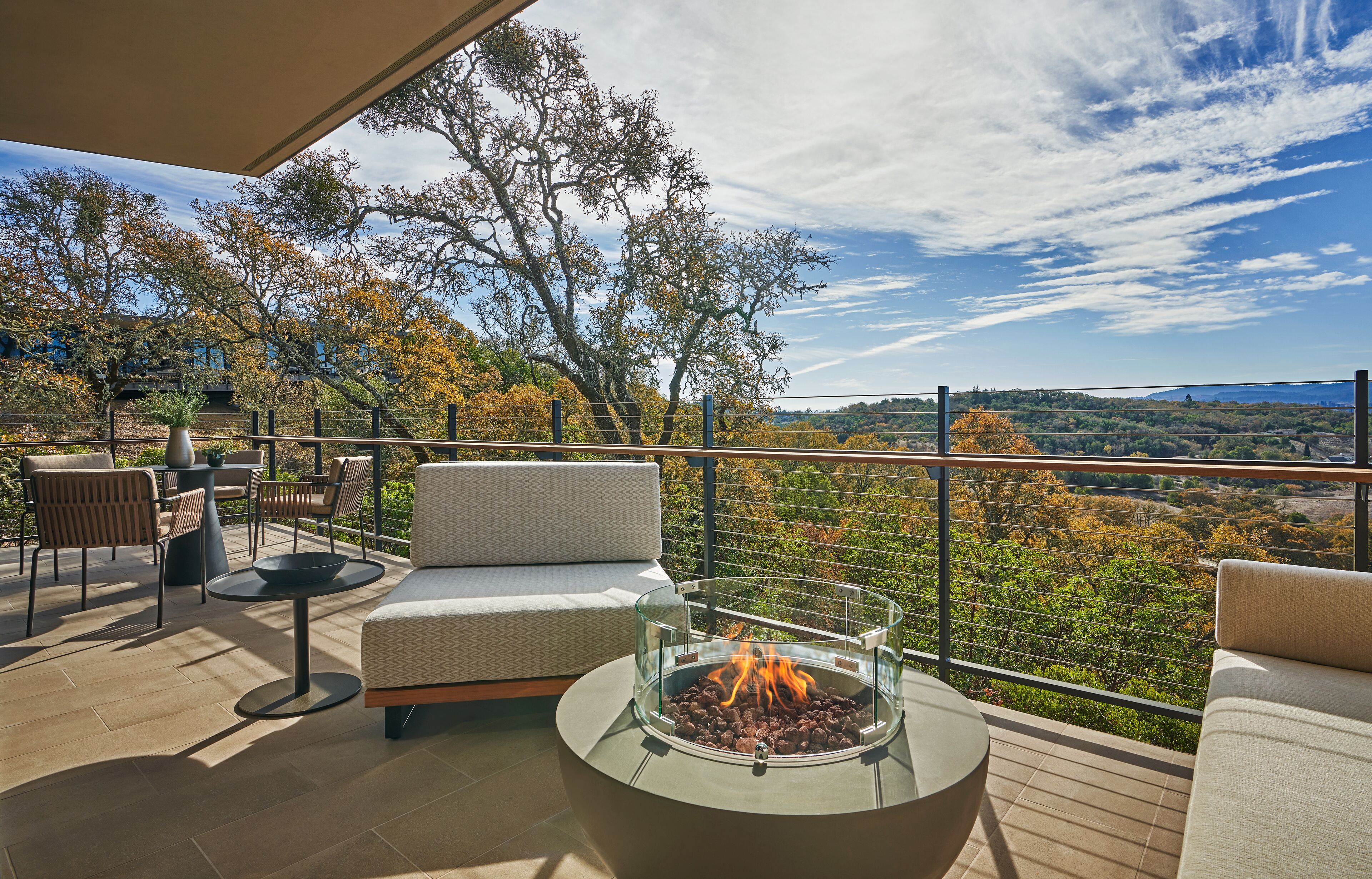 Una terraza con mobiliario exterior, un pozo de fuego y una vista panorámica de árboles y colinas.