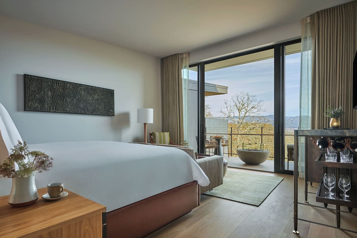 A modern hotel room with a large bed, a view of the outdoors, a dining area, and a bar with glasses.