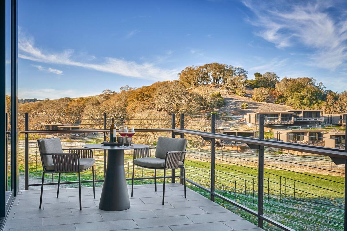 A balcony with two chairs and a table, overlooking a landscaped area with trees and buildings.