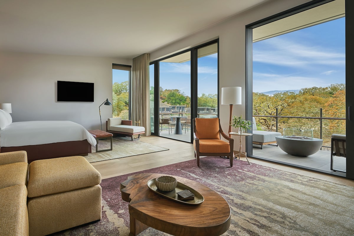 A modern hotel room with a large bed, a sofa, a chair, a small table, and a balcony with a view of trees and a distant landscape.