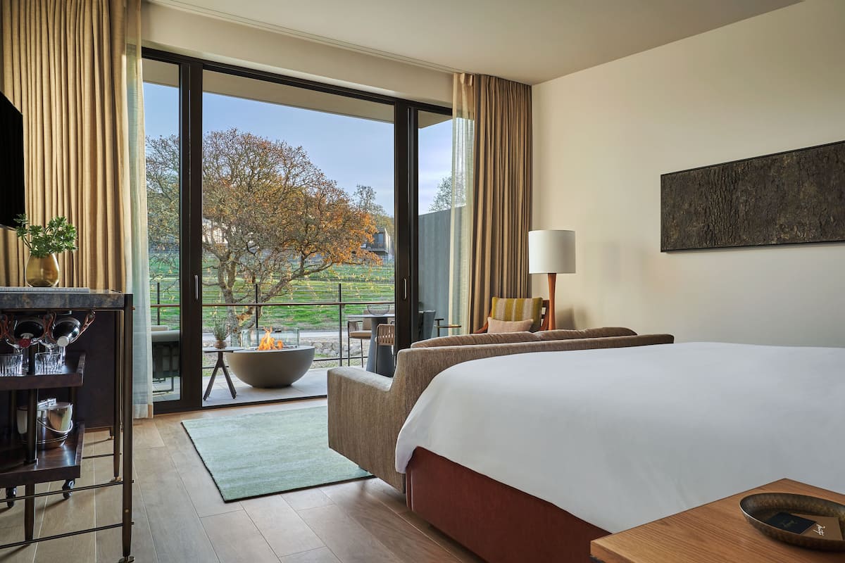 A hotel room with a large bed, a desk with a vase and books, a chair, a table with a bowl, and a view of trees and a building outside.
