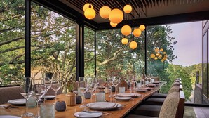A dining area with a long wooden table set for a meal, large windows offering a view of trees, and hanging spherical lights. - Montage Healdsburg (Healdsburg)