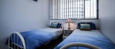 A small, clean room with two single beds, a bedside table with a lamp, and a window with vertical blinds.