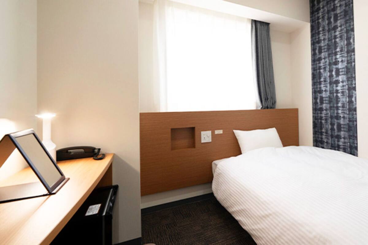 A hotel room with a wooden headboard, a bed with white bedding, a desk with a laptop, and a window with curtains.