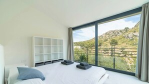 A modern bedroom with a large bed, a white shelving unit, and a view of a hilly landscape through a large window. - NATURE VILLA BONAIRE - Exclusive villa with infinity pool. (Alcúdia)