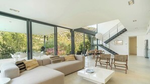 A modern living room with a large sectional sofa, a coffee table, and a dining area with chairs. There is a staircase leading to an upper level. - NATURE VILLA BONAIRE - Exclusive villa with infinity pool. (Alcúdia)
